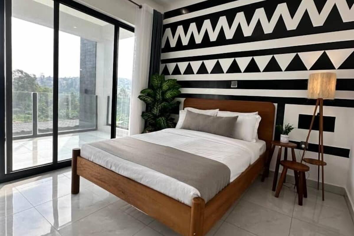 A modern bedroom with a large bed, a view through floor-to-ceiling windows, and a geometric wall design.