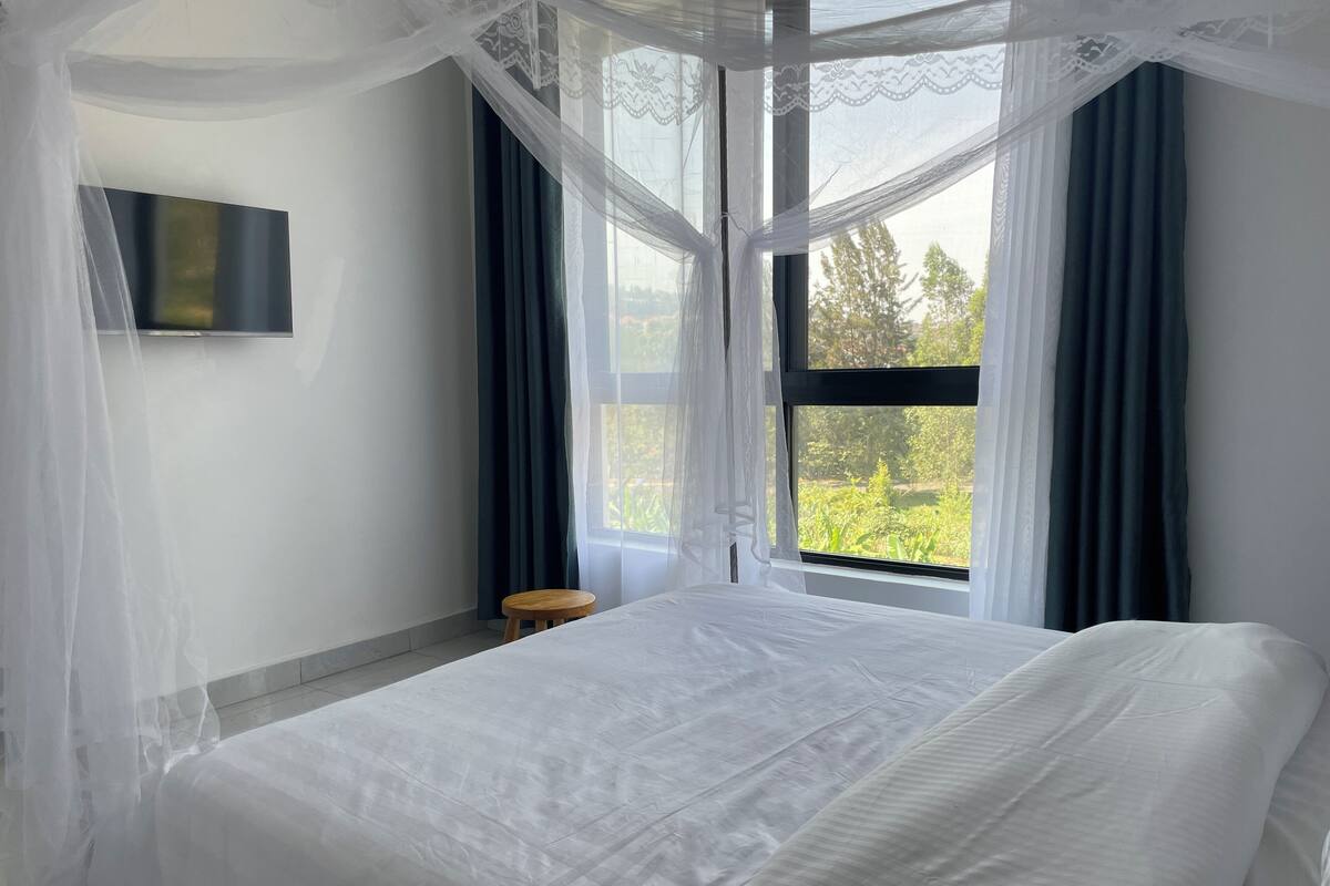 A bedroom with a large bed, a small wooden stool, a TV mounted on the wall, and a window with sheer curtains.