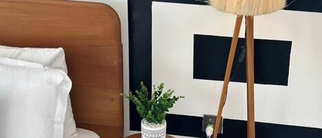 Un dormitorio con cama de madera, ropa de cama blanca, una pequeña mesa redonda de madera con una planta en maceta y una lámpara de pie alta con pantalla amarilla.
