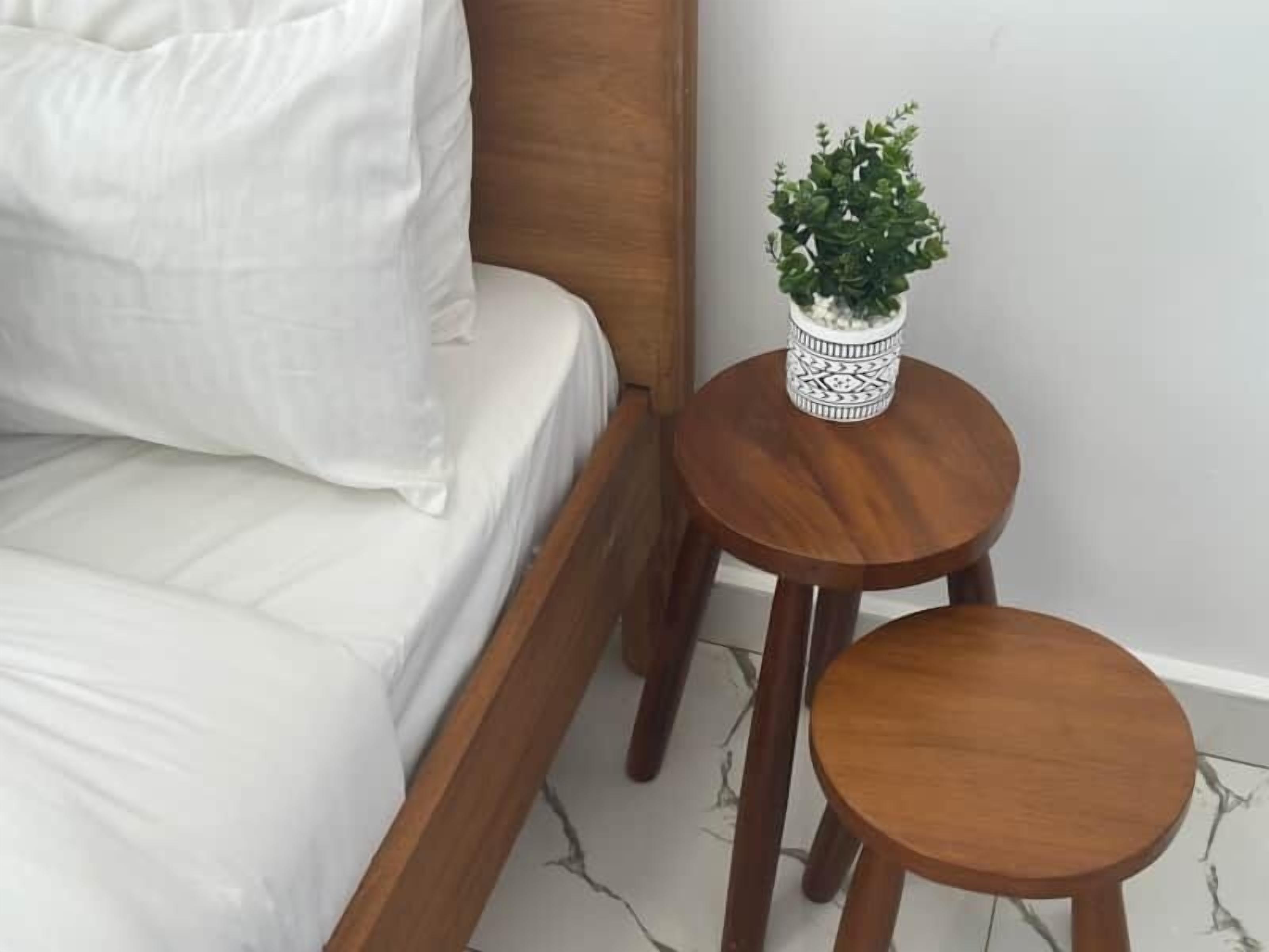A bed with white bedding, a wooden headboard, and a small wooden table with a potted plant beside it.