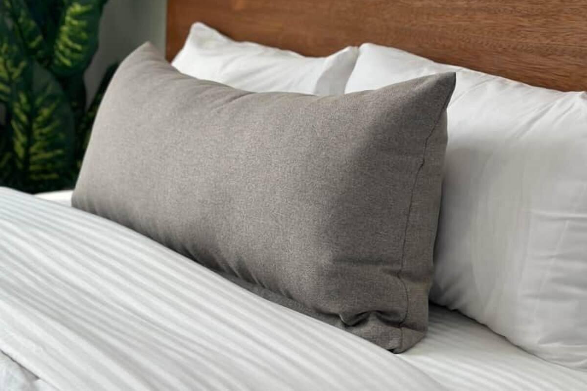 A neatly made bed with grey and white pillows, a wooden headboard, and a large green plant to the left.