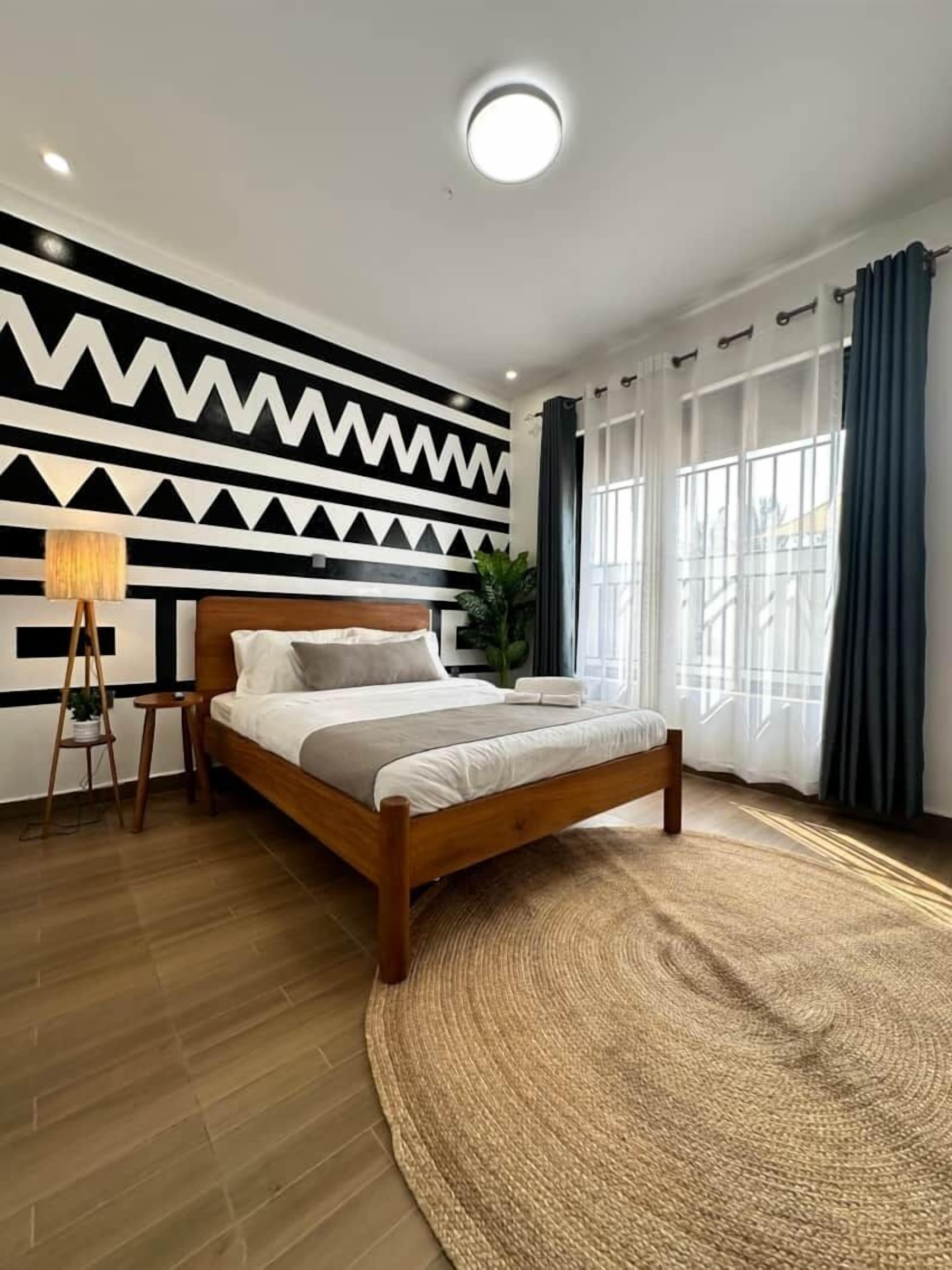 A modern bedroom with a wooden bed, a geometric wall design, a standing lamp, and large windows with curtains.