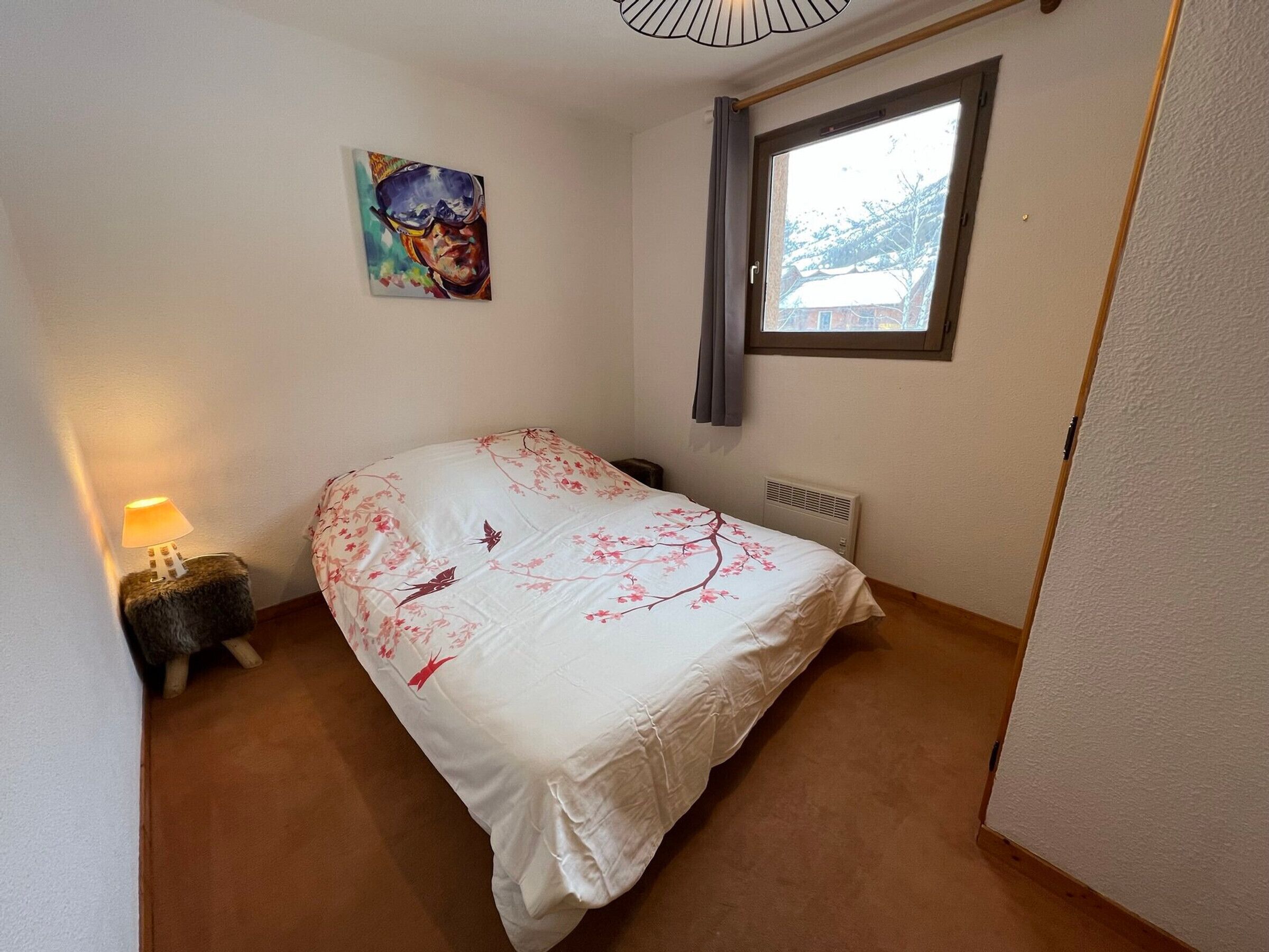 Un dormitorio con cama, mesita de noche, lámpara, radiador, ventana y un cuadro en la pared.