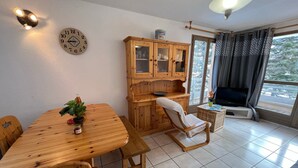 A cozy room with a wooden dining table, chairs, a wooden cabinet with glass doors, a TV, and a window with curtains. - Spacious apartment 8 guests, south-facing balcony, parking (La Salle-les-Alpes)