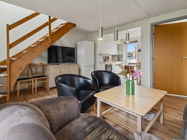 A modern living space with a wooden staircase, a flat-screen TV, a dining area with two chairs, and a kitchen in the background.