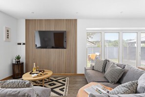 A modern living room with a flat-screen TV mounted on a wooden panel wall, a grey sofa with patterned cushions, a round wooden coffee table, and a large window offering a view of the outdoors.