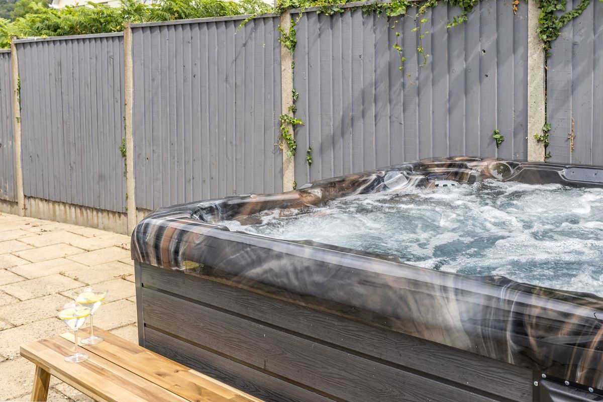 A hot tub with a wooden bench in front, surrounded by a grey fence with some greenery.