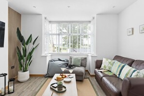 A modern living room with a brown leather sofa, a wooden floor, a large potted plant, and a window with white blinds.