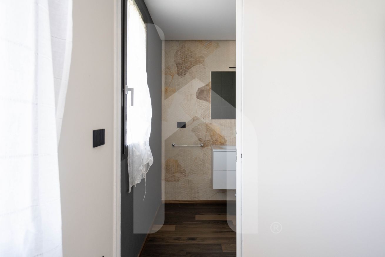 Un couloir étroit mène à une salle de bain dont le mur est orné d’un motif décoratif, qui contient également un porte-serviettes et un meuble blanc.