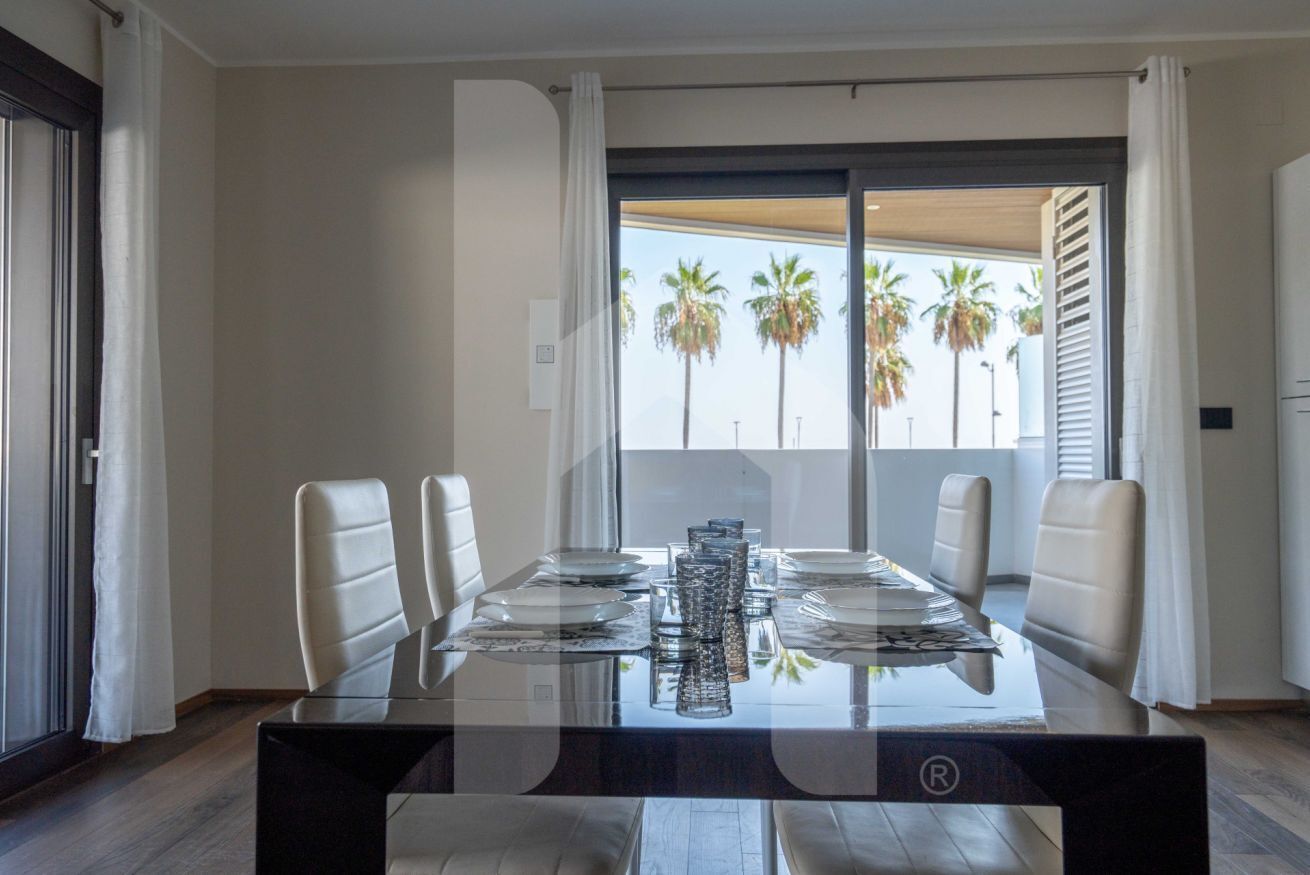 A modern dining area with a glass table, white chairs, and a view of palm trees through the balcony.
