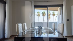 A modern dining area with a glass table, white chairs, and a view of palm trees through the balcony.