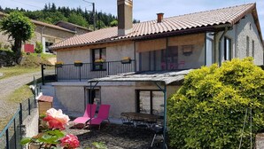 Une maison à deux étages avec une façade en pierre, un balcon agrémenté de chaises et un jardin où poussent des rosiers.