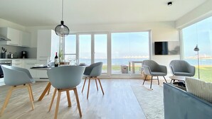 Cozinha moderna com armários brancos, área de jantar com cadeiras cinzas e uma grande janela com vista para o mar.
