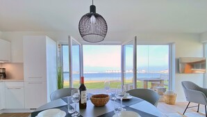 Uma sala de jantar com uma mesa posta para dois, uma garrafa de vinho e vista para o mar através de grandes janelas.