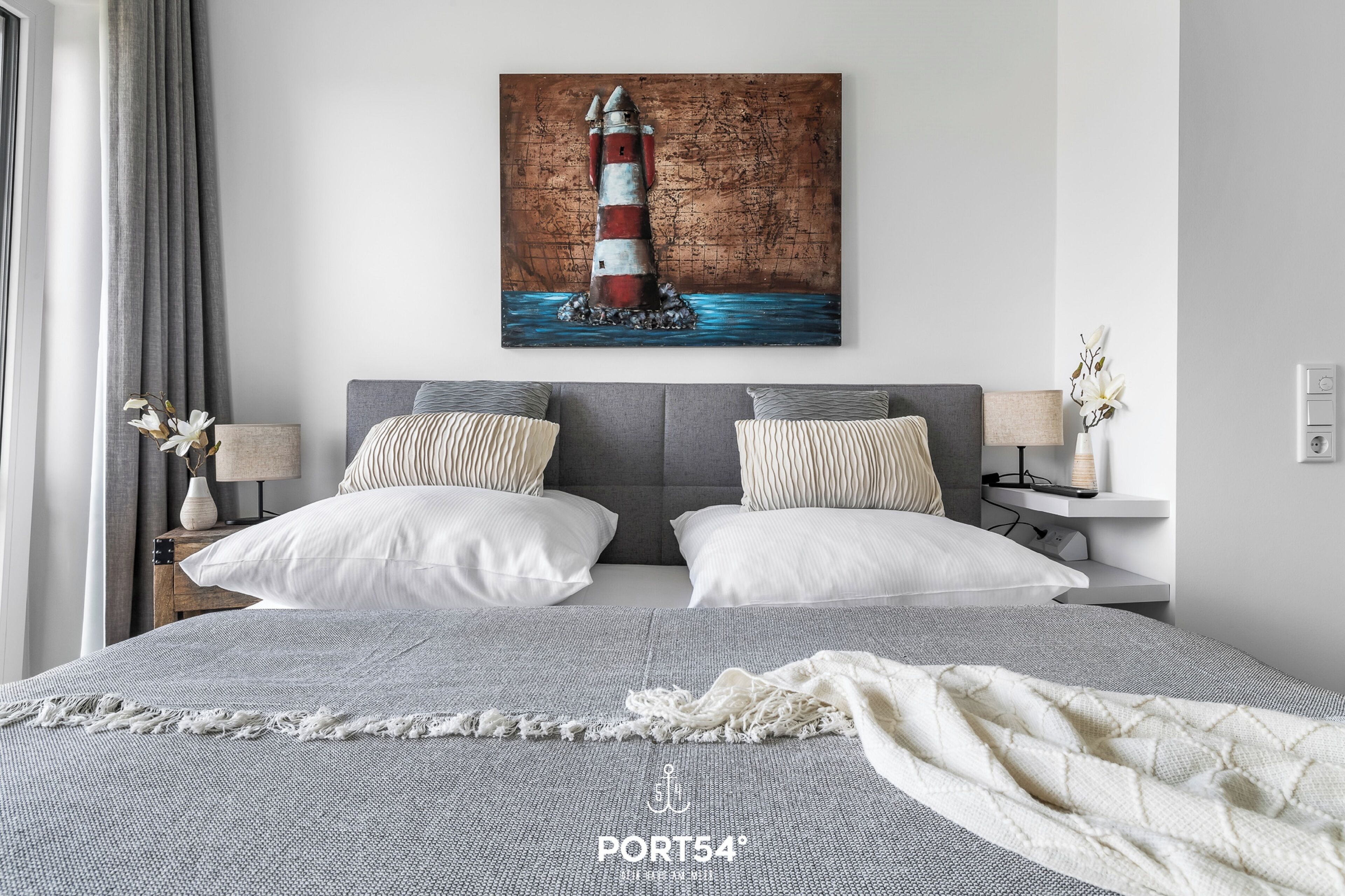 A modern bedroom with a large bed, two bedside tables with lamps, a painting of a lighthouse, and a view of the sea.