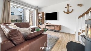 A modern living room with a brown leather sofa, a flat-screen TV mounted on the wall, and a wooden desk with a bookshelf. - Ferienhaus Wellenrauschen - Terrasse, Balkon, Sauna, Kamin, Nordsee- & Strandnah (Büsum)