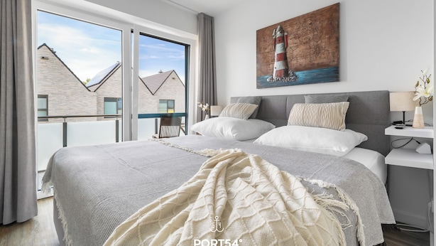 A modern bedroom with a large bed, a view of buildings outside, and a painting of a lighthouse. - Ferienhaus Wellenrauschen - Terrasse, Balkon, Sauna, Kamin, Nordsee- & Strandnah (Büsum)
