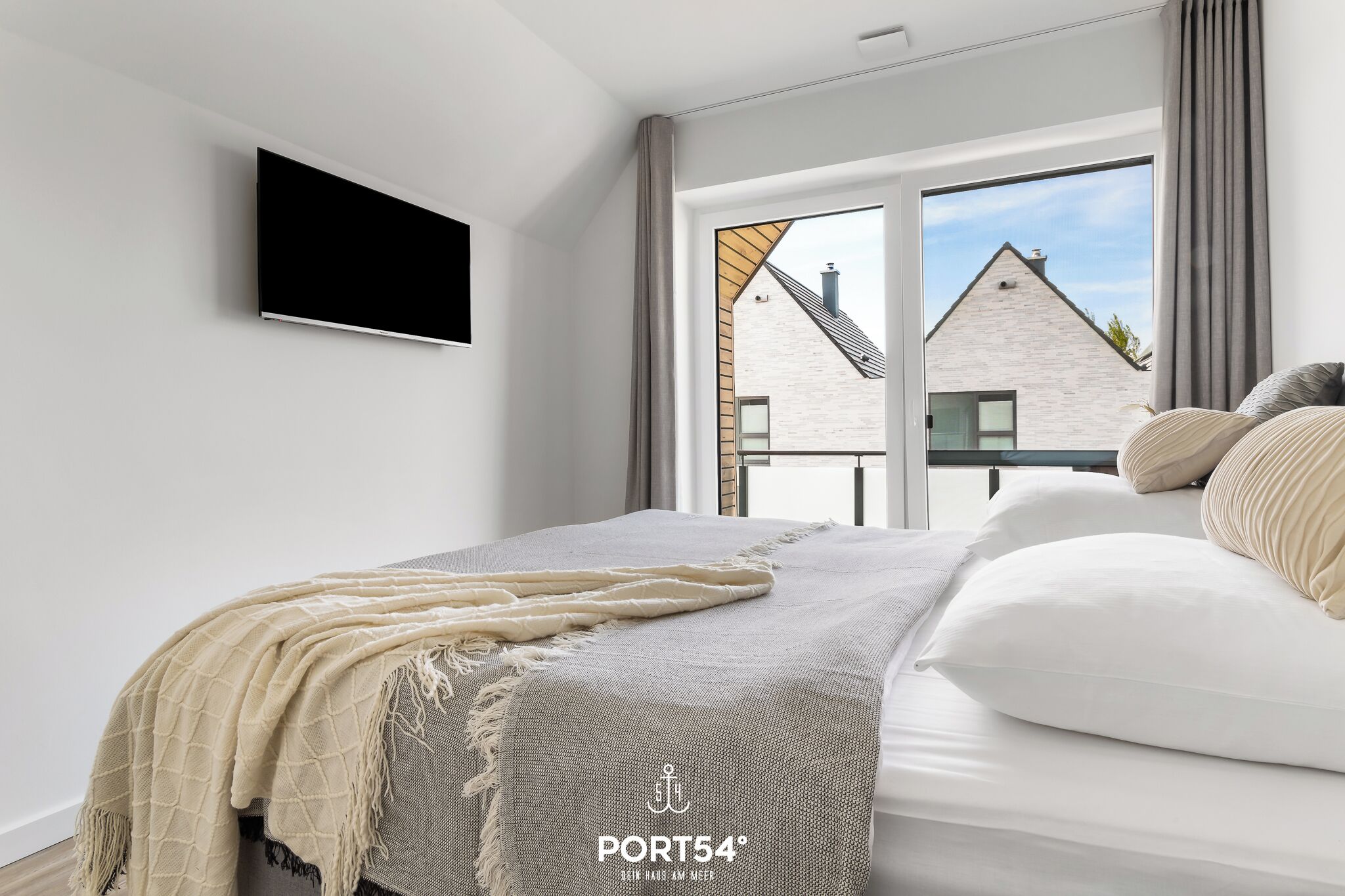 Une chambre moderne avec un lit, un téléviseur fixé au mur et une vue sur une maison par la fenêtre.