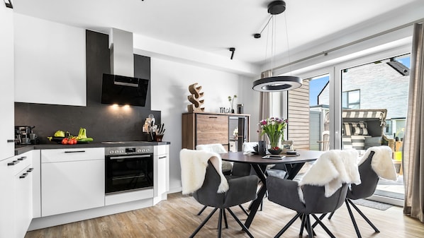 A modern kitchen with a built-in oven, a dining area with a round table and chairs, and a view of the outside. - Ferienhaus Wellenrauschen - Terrasse, Balkon, Sauna, Kamin, Nordsee- & Strandnah (Büsum)