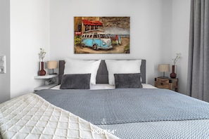 A bedroom with a bed, two bedside tables, a nightstand, a wall-mounted painting of a van, and a window with curtains. - Ferienhaus Wellenrauschen - Terrasse, Balkon, Sauna, Kamin, Nordsee- & Strandnah (Büsum)