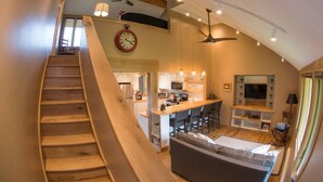 Ein geräumiger Wohnbereich mit einer Holztreppe, einer Küche im Hintergrund und einer gemütlichen Sitzecke mit Fernseher.