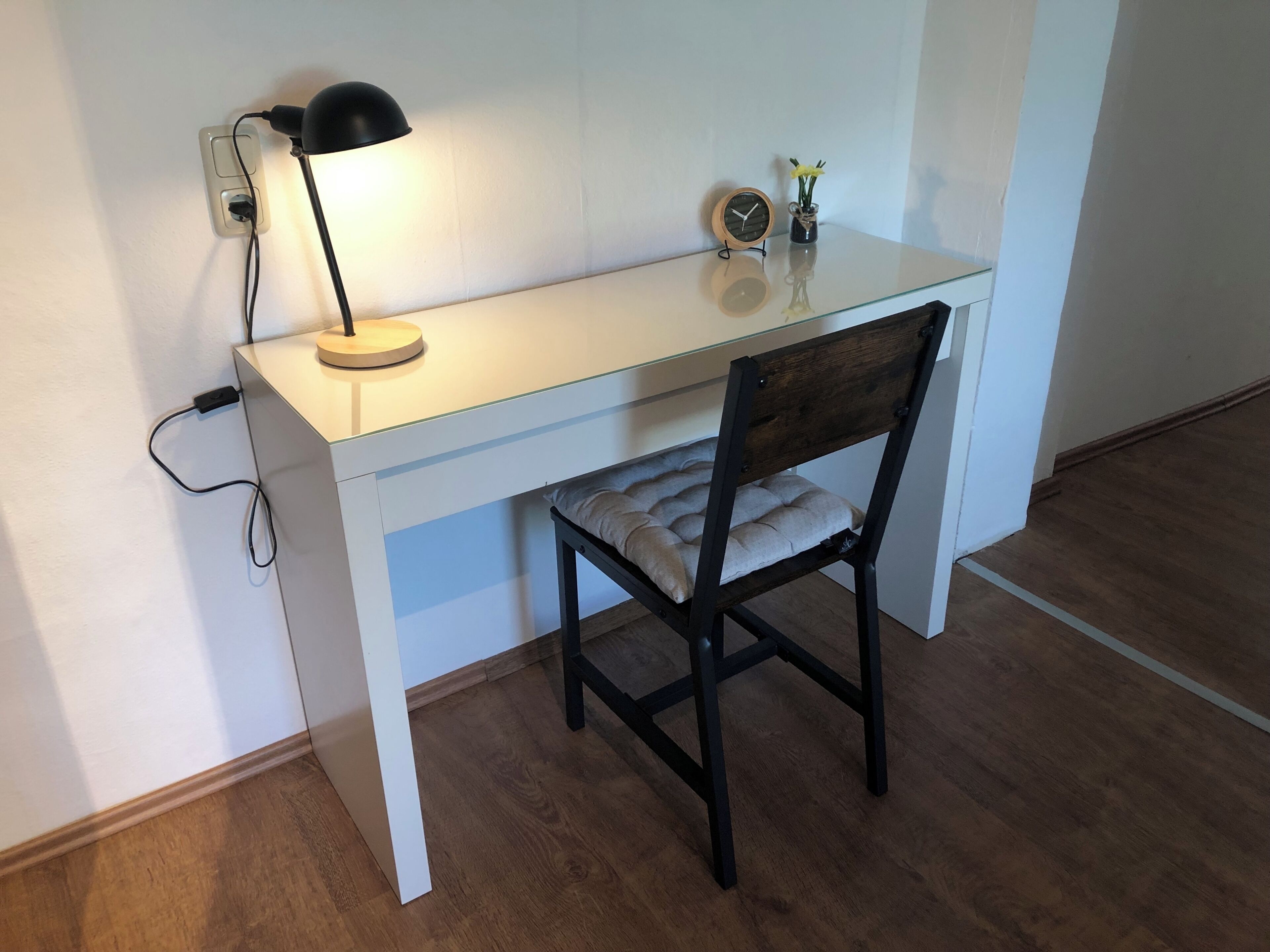 Un bureau blanc avec une lampe noire, une chaise en bois et une petite horloge au mur.