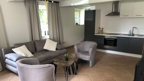 Ein modernes Wohnzimmer mit grauem Sofa, Sesseln und einem kleinen Couchtisch. Im Hintergrund ist eine Küche mit dunklen Schränken und einer Edelstahl-Dunstabzugshaube erkennbar.