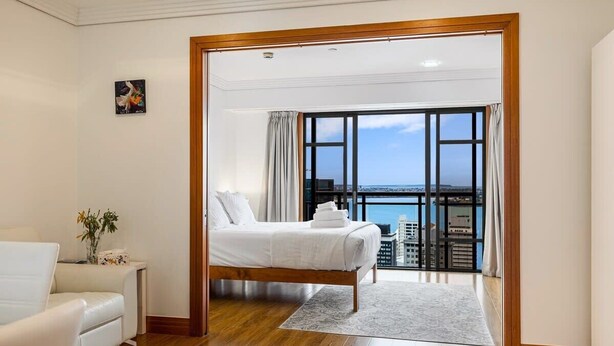 Une chambre à coucher comprenant un lit, une chaise, une table de chevet, une vue sur la ville et un balcon avec une balustrade.