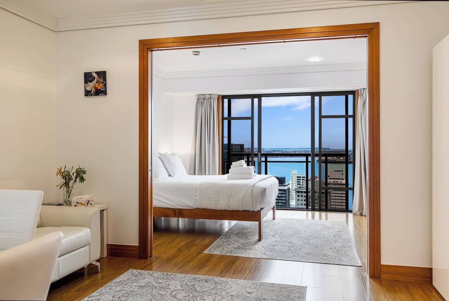 A hotel room with a bed, a white armchair, a view of the city, and a balcony.