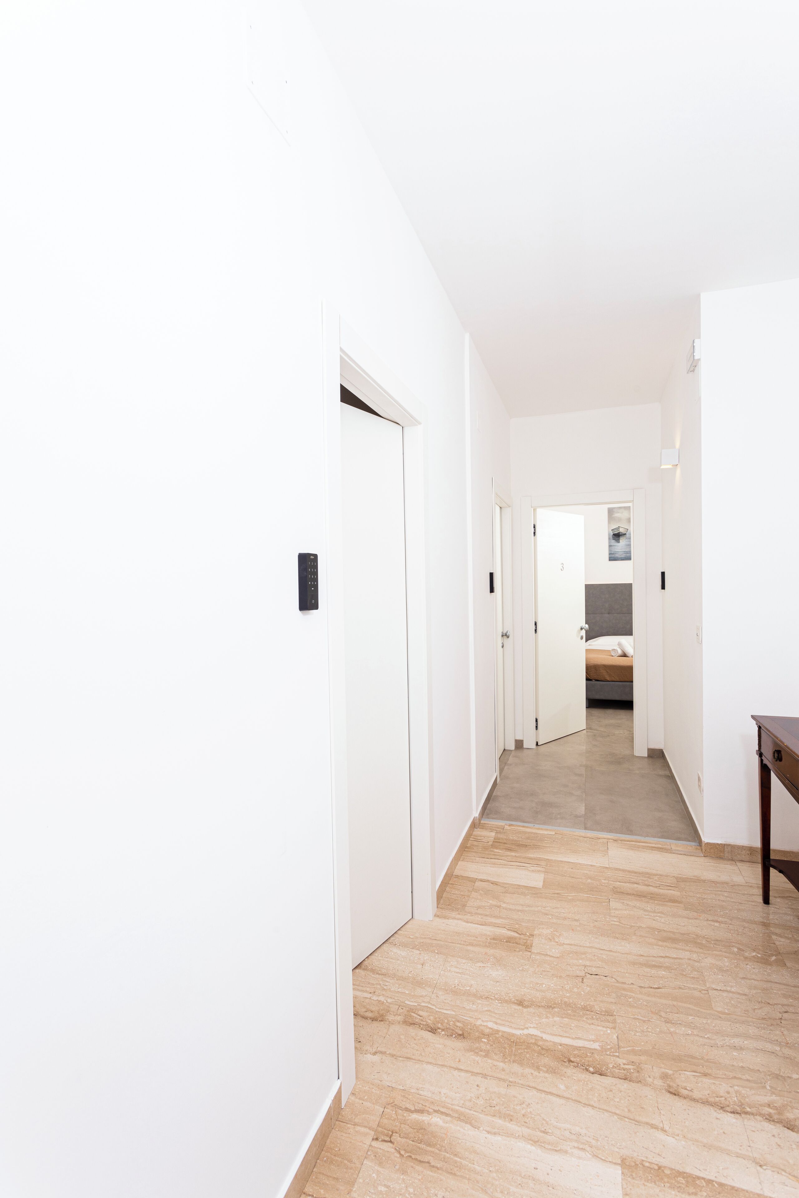 A narrow hallway with white walls and a wooden floor leading to a bedroom with a bed and a nightstand.