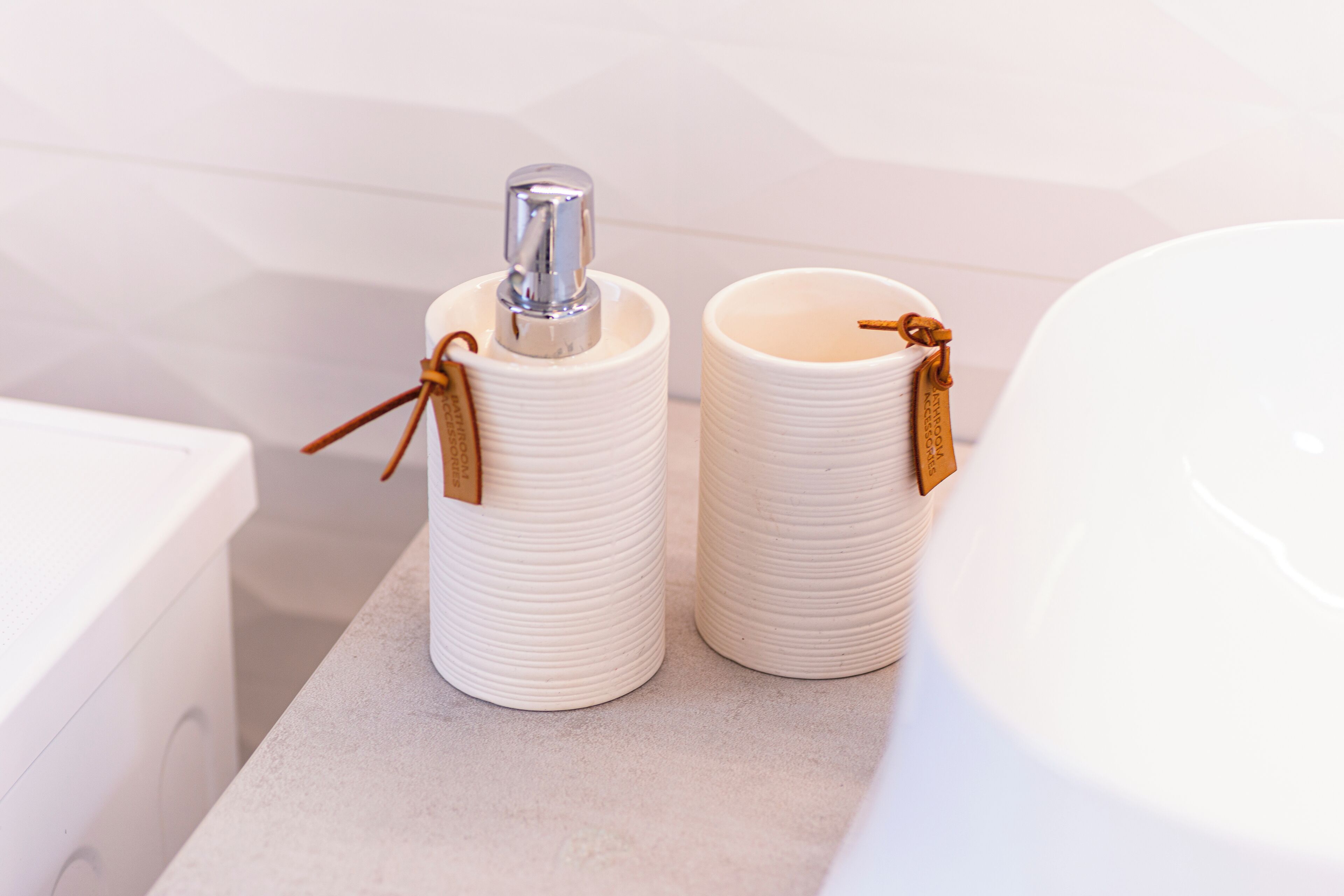 Two white ceramic containers with handles and tags on a bathroom countertop.