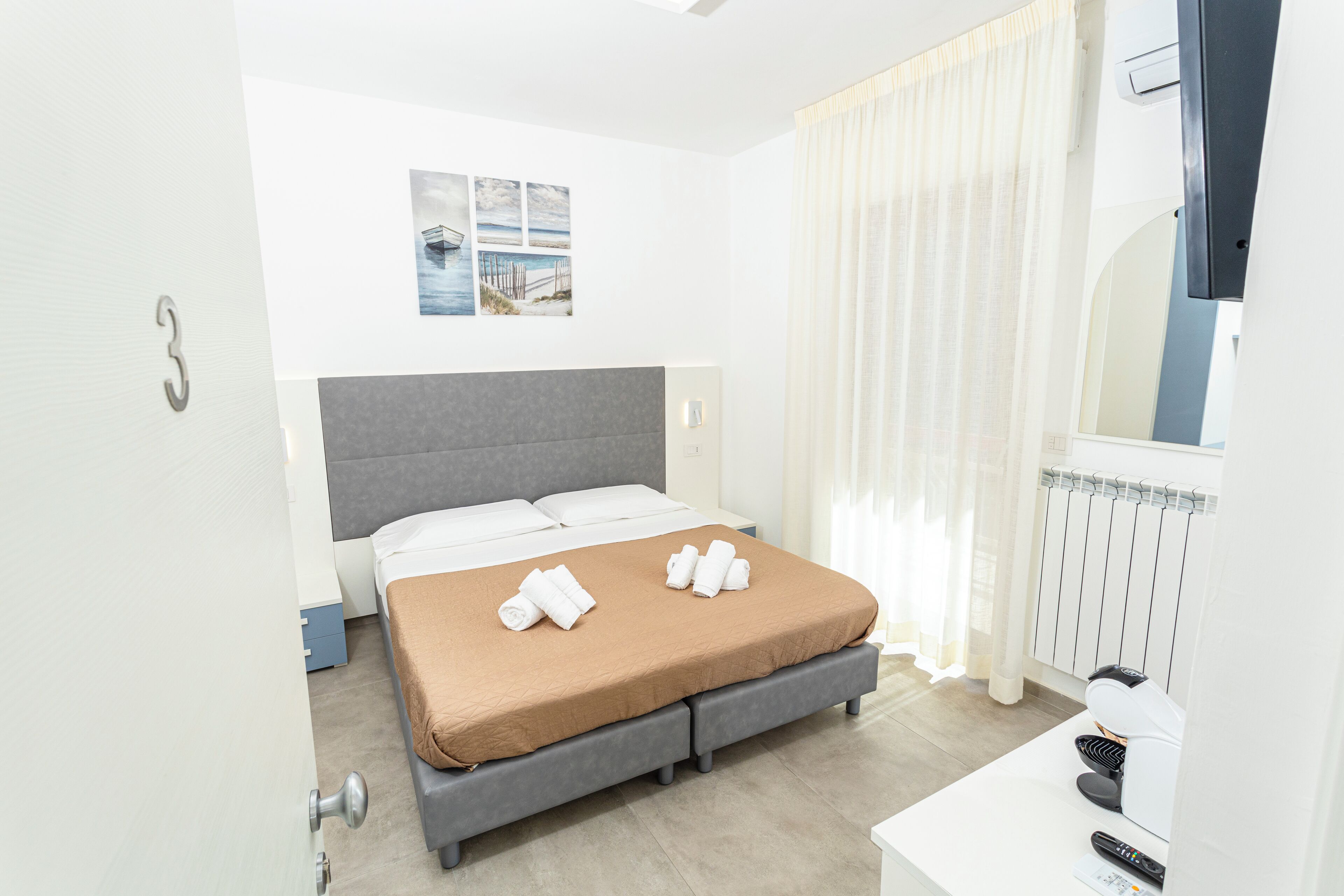 Moderni hotellihuone, jossa on sänky, yöpöytä, jossa on kahvinkeitin, peili ja ikkuna, jossa on verhot.
