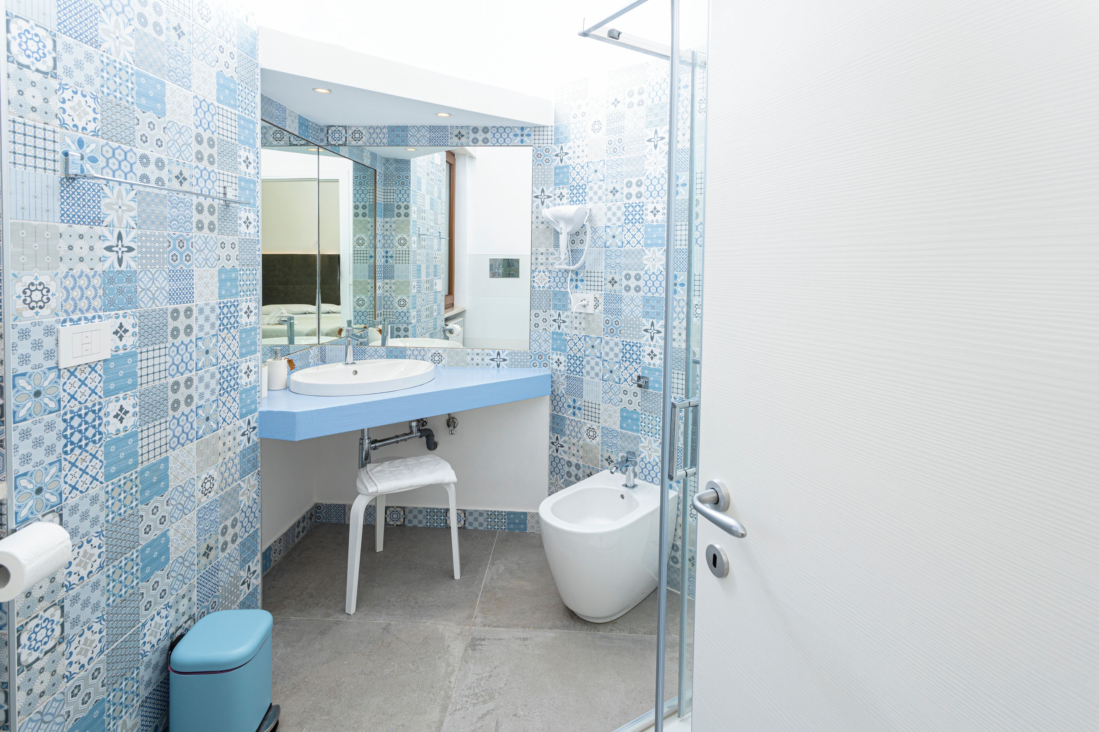 A bathroom with a blue and white tiled backsplash, a white sink, a mirror, a white chair, a bidet, and a glass shower enclosure.