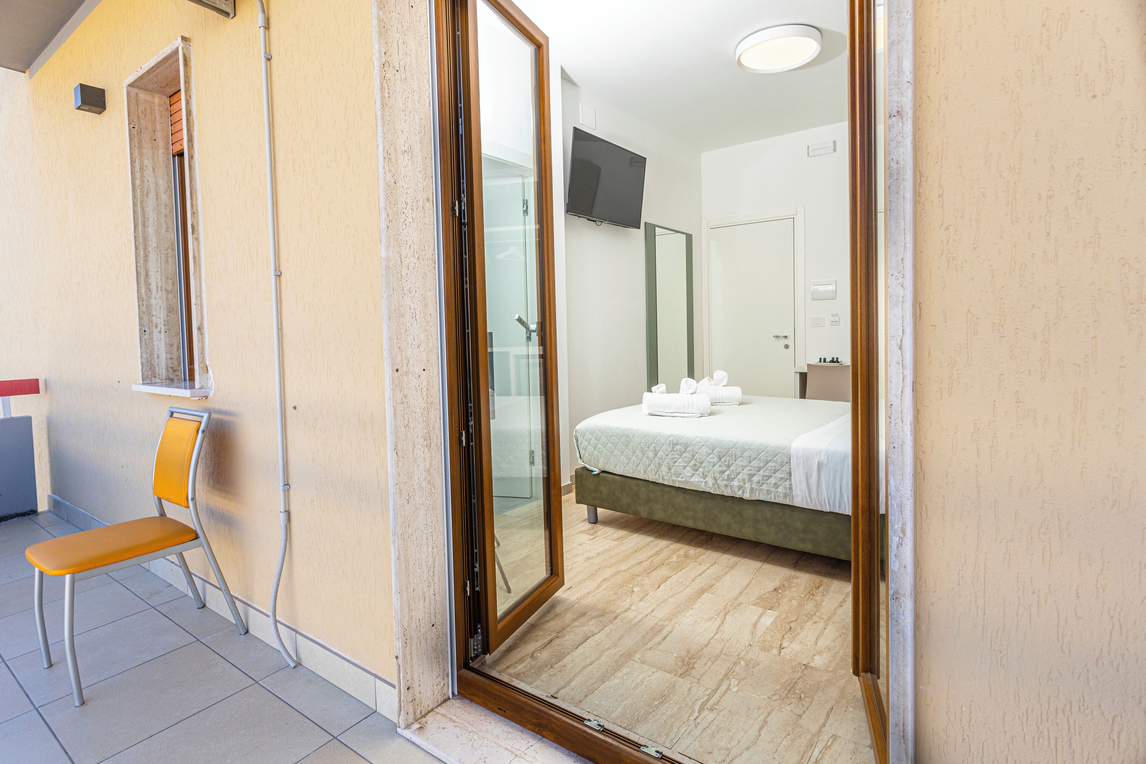Una camera d'albergo con un letto, una sedia, una TV e un bagno visibile attraverso una porta di vetro.