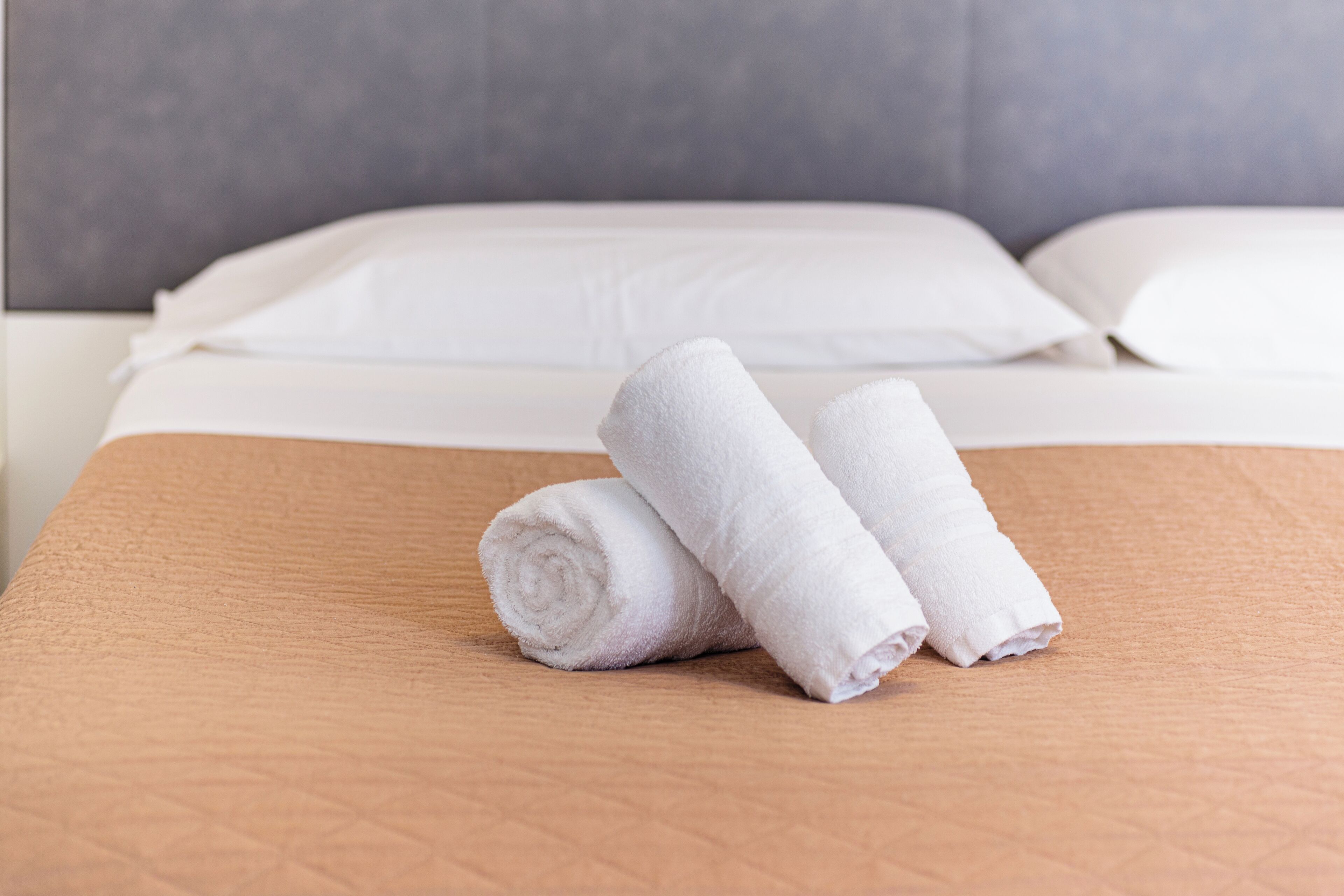 A neatly made bed with white linens and two rolled towels on a textured brown bedspread.