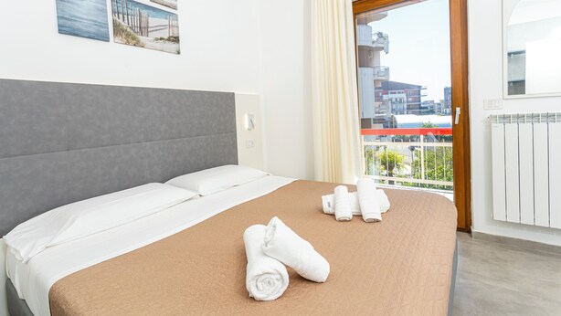 A hotel room with a bed, towels, a view of buildings, and a balcony. - Gallo's City Rooms (Termoli)