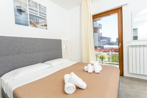 A hotel room with a bed, towels, a view of buildings, and a balcony. - Gallo's City Rooms (Termoli)