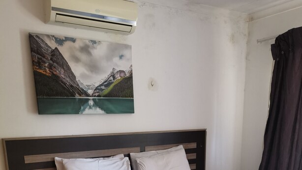 Una cama bien hecha con sábanas blancas y una manta turquesa, cabecera de madera oscura, una mesita de noche pequeña y un cuadro de un paisaje montañoso en la pared.