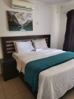 A neatly made bed with white linens and a teal throw blanket, a dark wooden headboard, a small bedside table, and a painting of a mountain landscape on the wall. - 97 High Road Guest house  (Edenvale)