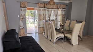 Una sala de estar con un sofá negro, una mesa de comedor de vidrio y cuatro sillas, y cortinas estampadas.