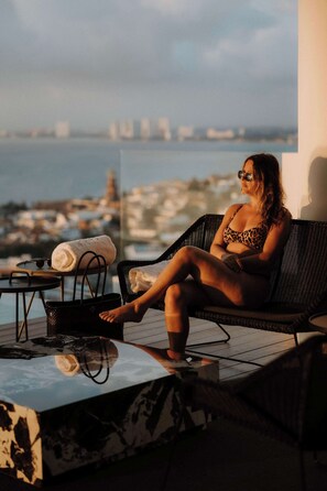 Eine Frau sitzt auf einer Korbschaukel mit Blick auf eine Stadtlandschaft und Wasser im Hintergrund.