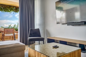 A modern living room with a glass coffee table, a grey upholstered chair, a wooden bench, and a large flat-screen TV mounted on the wall.