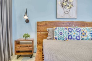 Une chambre à coucher avec une tête de lit en bois, des coussins à motifs, une table de chevet avec une plante et une lampe fixée au mur.