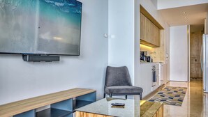 Un salon moderne avec un téléviseur à écran plat, un fauteuil gris, une table basse en bois et un coin cuisine à l’arrière-plan.