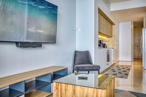 Un salon moderne avec un téléviseur à écran plat, un fauteuil gris, une table basse en bois et un coin cuisine à l’arrière-plan.