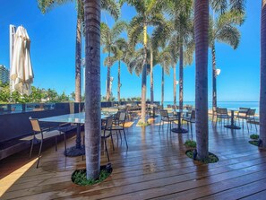 A rooftop bar with tables and chairs, palm trees, and a view of the ocean. - NEW Luxury Modern Condo in SOHO PV! Best Rooftop! (Puerto Vallarta)