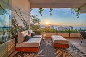 A balcony with a sofa and ottoman, a view of the sunset, and a cityscape in the background.