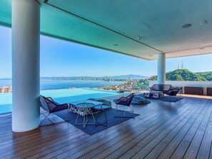 Un vaste espace extérieur doté d’une terrasse en bois, d’une piscine et de coins salon offrant une vue sur le paysage urbain de la côte.