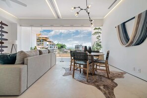 Una sala de estar moderna con sofá, mesa de comedor, sillas y vista a edificios a través de amplias ventanas.