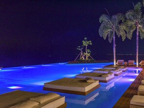 Una piscina infinita con camas flotantes y palmeras, rodeada de sillones de descanso.